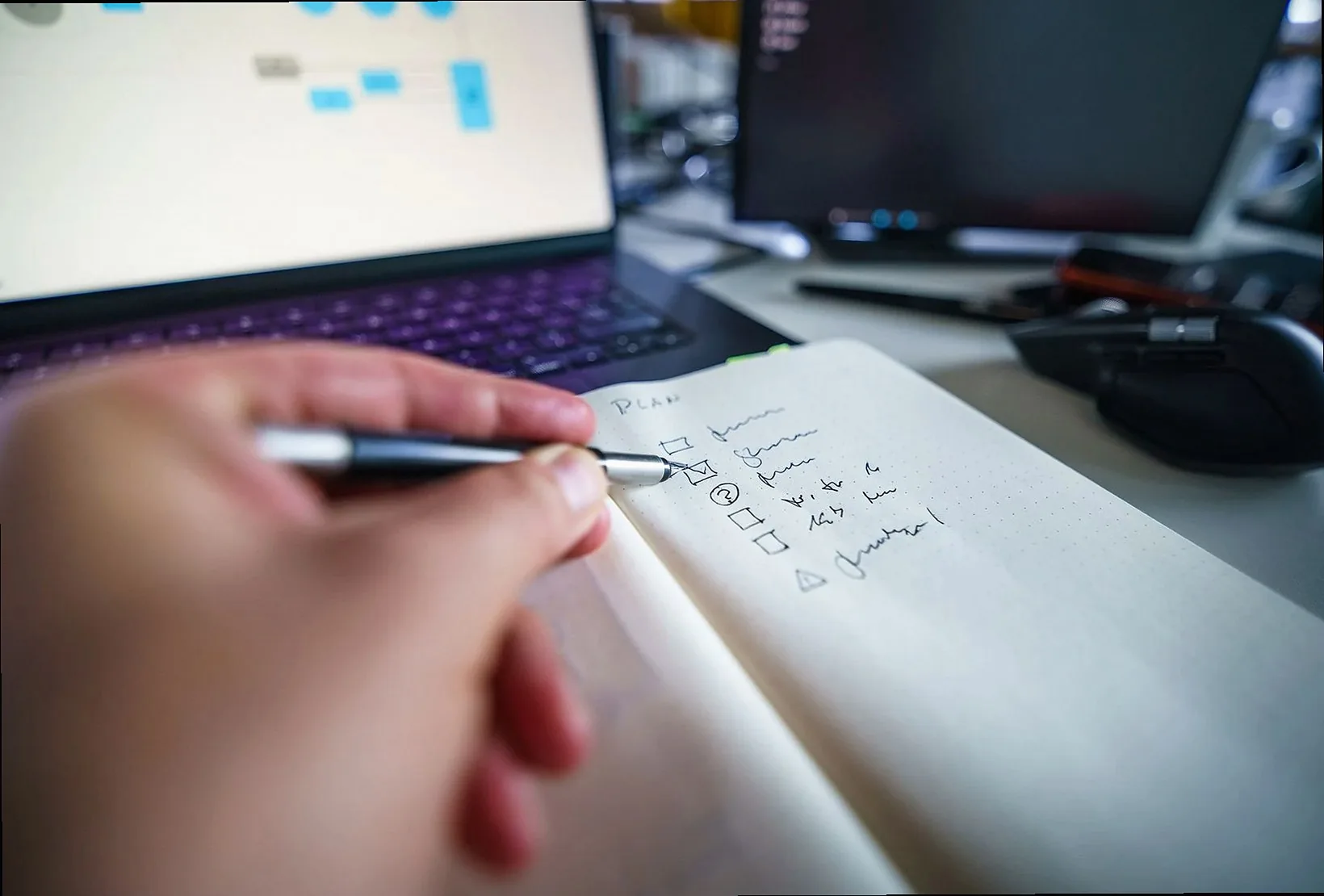 ノートパソコンのあるデスクでノートに手書きし、生産性と整理整頓のスキルを示している。